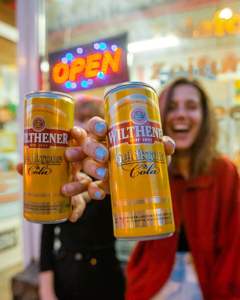 Zwei Personen präsentieren lachend Dosen von “WILTHENER Goldkrone Cola” vor einem leuchtenden “OPEN”-Schild an einem Kiosk- oder Späti-Ladeneingang.