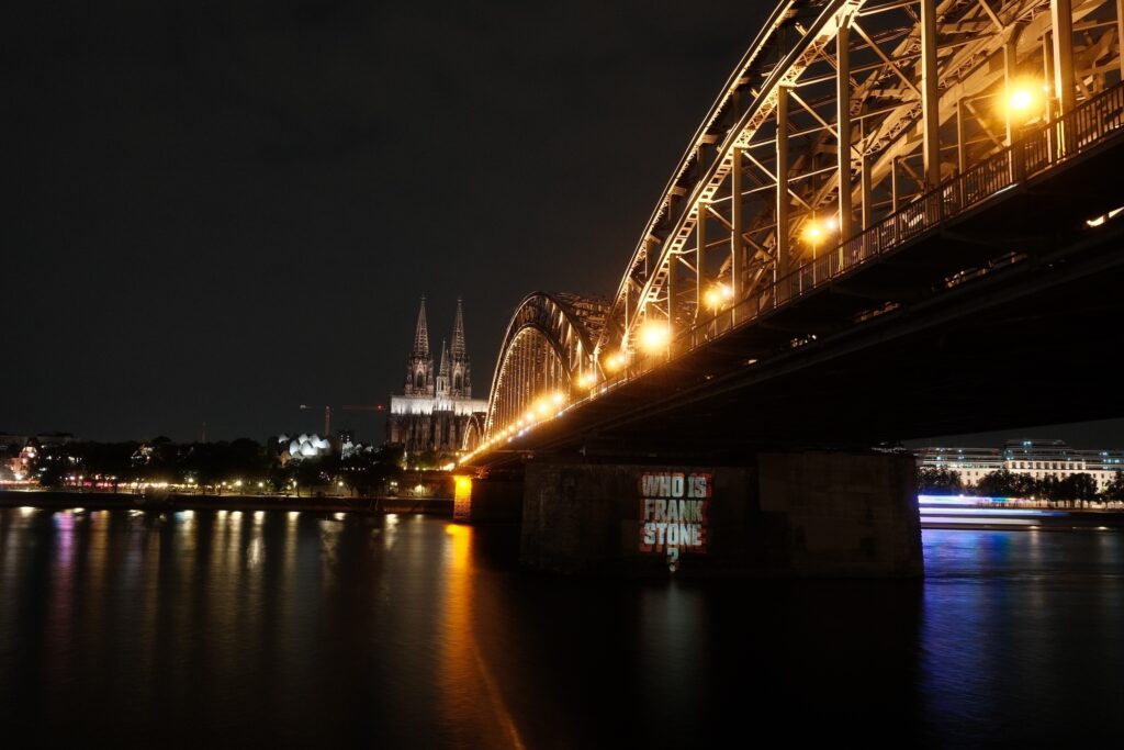 Kölner Stadtpanorama bei Nacht. Auf einem Brückenpfeiler der Hohenzollernbrücke ist eine Lichtprojektion mit der Aufschrift "Who is Frank Stone?" zu sehen. Im Hintergrund ist der Kölner Dom zu erkennen.