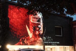 Projektion einer Bildszene aus dem Horrorspiel "The Casting of Frank Stone" auf einem Gebäude. Es ist ein Mann mit einer Eisenmaske aus der ein Auge und Mund und Kiefer sichtbar sind.